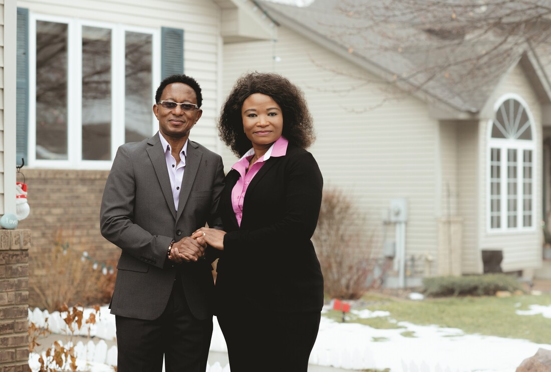 Edwin Swaray stands on the left with his wife, Vivian Ballah-Swaray on the right, in front of a series of houses.