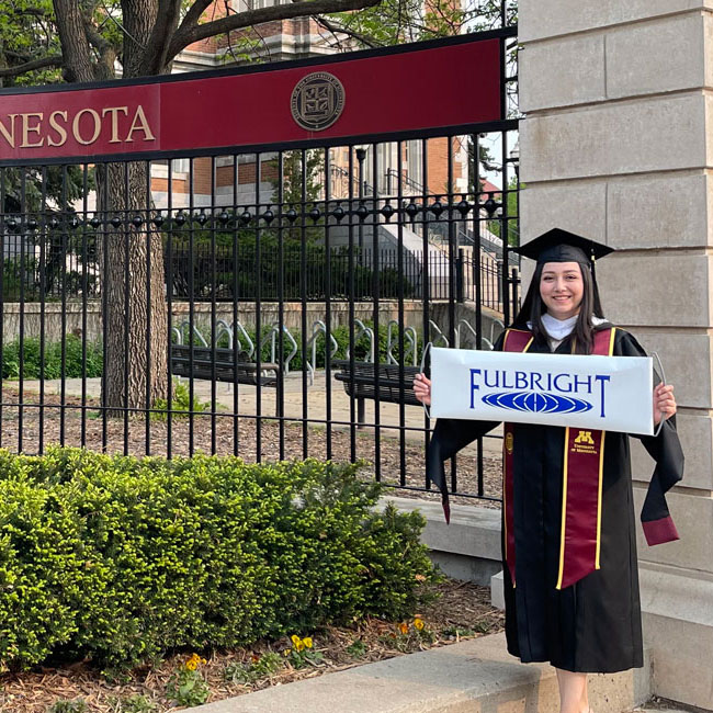 Student in graduation robes holding a Fulbright banner in front of a fence with the University of Minnesota wordmark