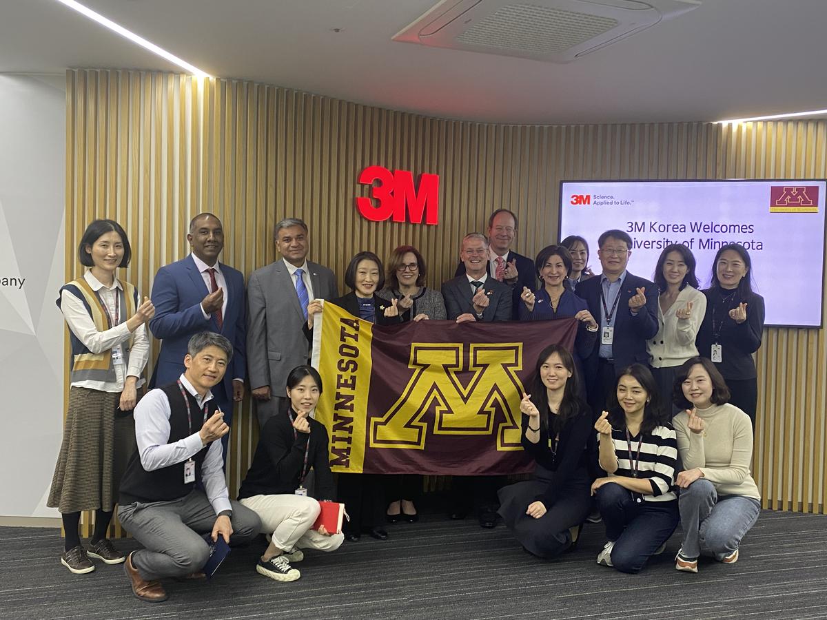 UMN delegation poses with M flag in front of wall with 3M logo