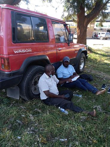 Nick sitting on the ground with another man, leaning against a red vehicle