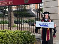Student in graduation robes holding a Fulbright banner in front of a fence with the University of Minnesota wordmark