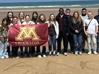 Students pose with a University of Minnesota Crookston flag on a beach in France